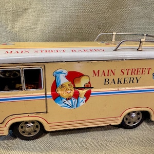 May include: Vintage toy bakery truck in cream and white with red and blue accents. The truck features the text "MAIN STREET BAKERY" and a graphic of a chef holding bread. A Dalmatian dog graphic is on the side.