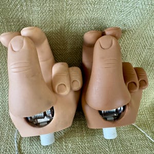 May include: Two light-toned plastic hand-shaped objects with fingers positioned in a crossed gesture. Each hand features a visible metal hinge mechanism at the wrist area and a white threaded plastic screw base extending from the bottom of the wrist.