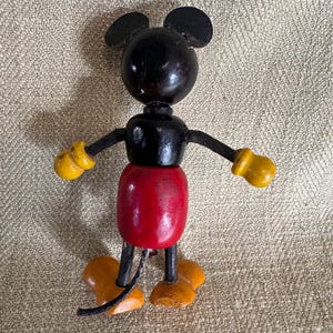 Puede incluir: Una figura de madera vintage de Mickey Mouse. La figura tiene una cabeza negra con orejas redondas, un cuerpo negro, un torso rojo, guantes amarillos y zapatos naranjas. Los brazos y las piernas son articulados.
