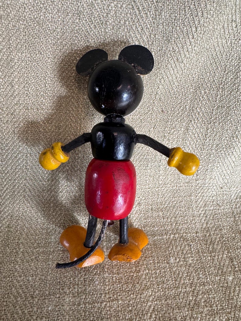 Puede incluir: Una figura de madera vintage de Mickey Mouse. La figura tiene una cabeza negra con orejas redondas, un cuerpo negro, un torso rojo, guantes amarillos y zapatos naranjas. Los brazos y las piernas son articulados.