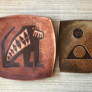 May include: Two ceramic plates with abstract designs. The first plate is brown with a black and white geometric animal design. The second plate is brown and green with a circle and triangle design.