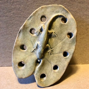 May include: A ceramic wall hanging in the shape of a leaf with a lizard sculpted on top. The leaf has multiple holes around the edge.
