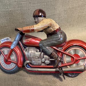 May include: Vintage red and silver toy motorcycle with a rider wearing a brown helmet and goggles. The motorcycle has a detailed engine and exhaust pipes. The word "Technofix" is on a small sign on the front.