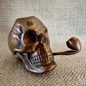 May include: A bronze-colored skull-shaped pipe with a detailed design. The pipe's bowl extends from the skull's mouth. The skull has visible teeth and eye sockets. The overall aesthetic is antique and gothic.
