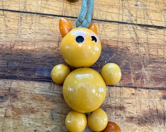 May include: A yellow wooden toy with a string attached to the top. The toy is shaped like a person with a round head and large eyes. The toy is made of multiple wooden beads.