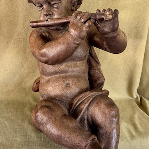 May include: A detailed wooden sculpture of a cherub playing a flute. The cherub is seated, with detailed carving of the hair, facial features, and body. The sculpture is a warm brown color, with visible wood grain.
