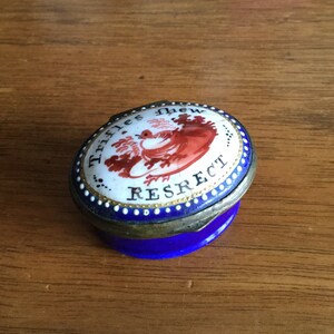May include: A small, oval-shaped, blue and white porcelain trinket box with a gold trim. The lid features a red bird with the words "Trifles Light" and "RESPECT" written around it.