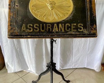 May include: A vintage wooden sign with a gold sun and the text "Cie du SOLEIL ASSURANCES" on a black background. The sign is mounted on a black metal tripod stand.
