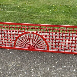 May include: A red metal grate with a decorative design. The grate has a repeating pattern of squares and a sunburst design in the center.