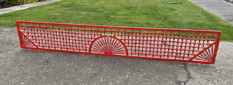 May include: A red metal grate with a decorative design. The grate has a repeating pattern of squares and a sunburst design in the center.