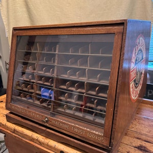May include: A wooden display case with glass doors and shelves for storing spools of thread. The case has a label that reads "D.W.HURST'S SYLKO' MACHINE TWIST 100 YDS."