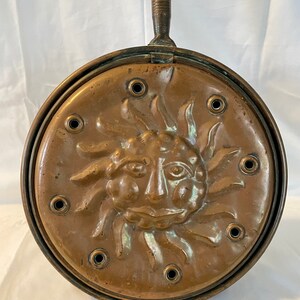 May include: Antique copper warming pan with a sun face design and a wooden handle. The pan has multiple holes around the sun face.