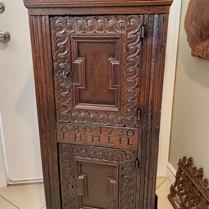 May include: A dark brown wooden cabinet with two doors and intricate carvings. The top door has a carved design and the bottom door has a carved design with a keyhole. The cabinet has a date carved on the front: "ANO 1730 I.H.M.E.L."
