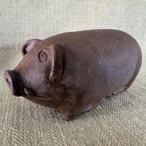 May include: A brown ceramic pig figurine with a rounded body, small ears, and a prominent snout. The pig has a rustic, aged appearance, suggesting it may be vintage or handcrafted. The figurine is set against a textured, light green background.