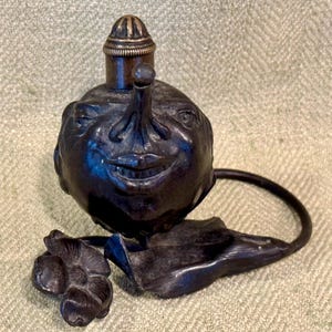 May include: A black metal bottle stopper shaped like a face with a large nose and a wide smile. The stopper has a small, round, gold-colored top and a ring handle. The stopper is sitting on a black metal base with a flower-shaped design.