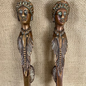 May include: Two antique bronze-toned metal decorative pieces. Each features a detailed face with a headdress and a stylized feather design. The pieces have a vintage aesthetic and are likely decorative elements.