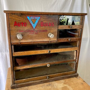 May include: A vintage wooden display case with glass doors and drawers. The case is labeled "CLASCO CANADIAN BULBS AUTO" and has a red triangle logo with the text "CLASCO CANADIAN BULBS".