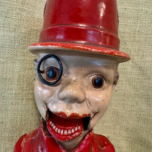 May include: A vintage, novelty bank in the shape of a man's head. The bank is painted with a red top hat and suit, with a white face, a monocle, and a wide, open mouth revealing teeth. The bank has a distressed, antique appearance.