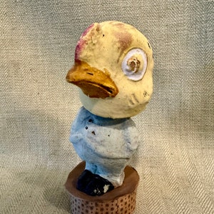 May include: A ceramic figurine of a bird with a yellow head, blue body, and brown base. The bird has a single eye and a beak.