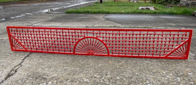 May include: A red metal grate with a decorative design featuring a sunburst pattern. The grate is made of a series of small, evenly spaced squares.