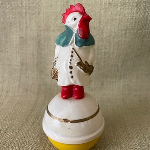 May include: A white ceramic rooster wearing a blue and green coat and red boots, standing on a yellow and white roly-poly toy.