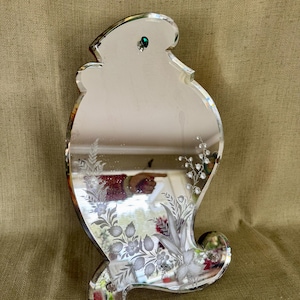 May include: A decorative, clear glass mirror with an ornate, etched floral design. The mirror has a unique, curved shape and a small, green jewel at the top. The etched design features flowers and leaves.