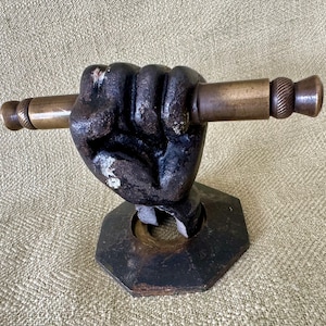 Puede incluir: Una mano de hierro fundido sosteniendo una varilla de latón con una perilla decorativa. La mano está cerrada en un puño y está montada sobre una base octogonal negra.