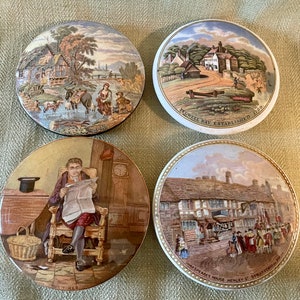 May include: Four antique ceramic lidded jar tops with colorful painted scenes. The tops feature a man reading a newspaper, a building with a sign that reads "Magpie's House Henley & Stratford R.R.", a building with a sign that reads "Powell Bay Established 1800", and a rural scene with a man and woman walking.