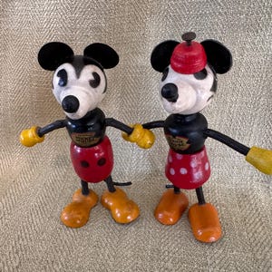Puede incluir: Dos figuras de madera vintage de Mickey Mouse y Minnie Mouse. Mickey tiene un cuerpo negro, pantalones cortos rojos y guantes y zapatos amarillos. Minnie tiene una falda roja con lunares blancos y un sombrero rojo. Ambos tienen orejas negras.