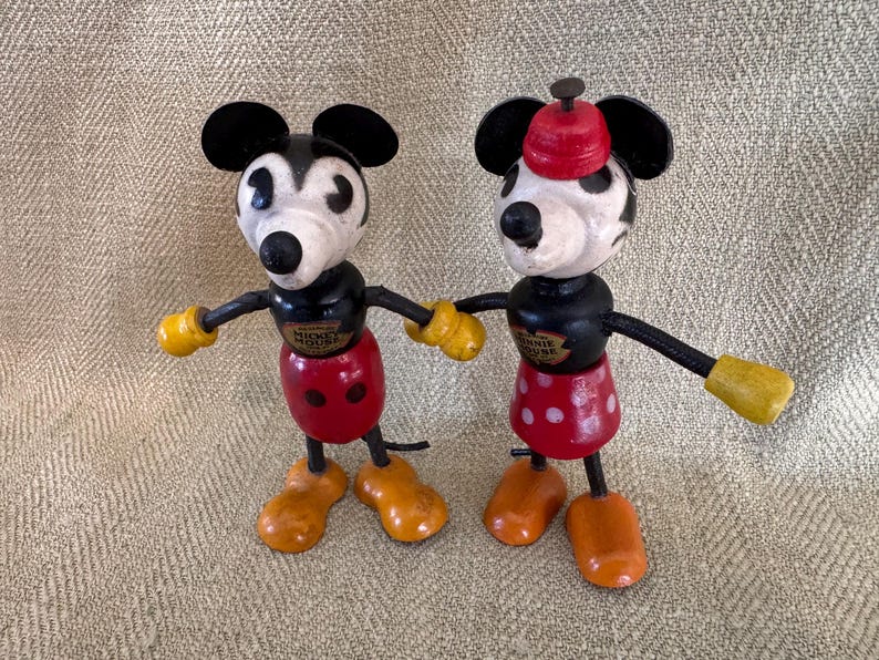 Puede incluir: Dos figuras de madera vintage de Mickey Mouse y Minnie Mouse. Mickey tiene un cuerpo negro, pantalones cortos rojos y guantes y zapatos amarillos. Minnie tiene una falda roja con lunares blancos y un sombrero rojo. Ambos tienen orejas negras.
