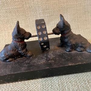 May include: Black cast iron figurine of two Scottish Terriers pulling a black and white wheel. The figurine is on a black base.