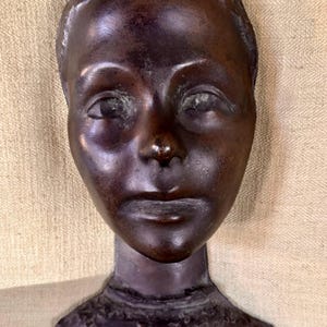 May include: A bronze sculpture of a human face, featuring detailed facial features. The sculpture has a dark brown patina and is mounted on a neck and base with ornate detailing. The face has a serene expression.