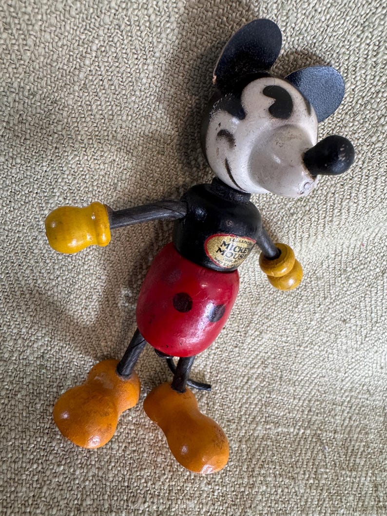 Puede incluir: Juguete de madera vintage de Mickey Mouse con orejas negras, cara blanca y nariz negra. La figura tiene un torso negro, pantalones cortos rojos y guantes y zapatos amarillos. Las palabras "Mickey Mouse" son visibles en el pecho.