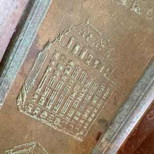 May include: Close-up of a copper-colored metal tray with embossed architectural designs. The tray features detailed depictions of buildings, possibly historical landmarks, with a textured surface and a raised border.