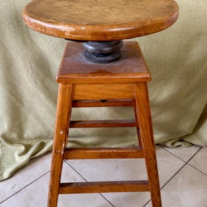 May include: A wooden artist's stool with a swiveling top. The stool has four legs and a rectangular base. The top is made of wood and has a smooth, rounded surface.