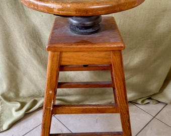 May include: A wooden artist's stool with a swiveling top. The stool has four legs and a rectangular base. The top is made of wood and has a smooth, rounded surface.