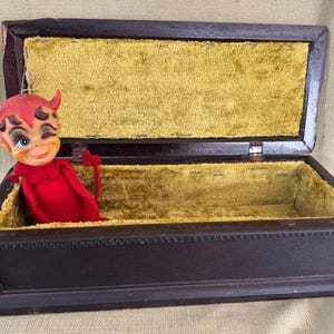 May include: A vintage wooden box with a hinged lid, lined with gold velvet. A red devil figurine with horns and a mischievous expression is inside. The box is dark brown.