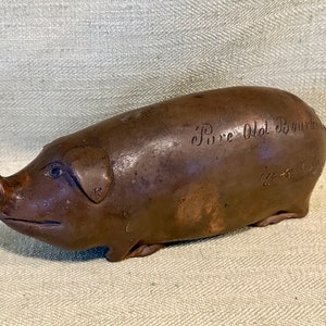 May include: A brown ceramic pig piggy bank with a snout that is open for inserting coins. The pig has a handwritten inscription on its side that reads "Pure Old Bourbon".