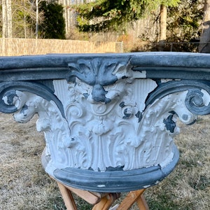 May include: A vintage, white and gray, plaster, decorative column capital with intricate floral and scrollwork details. The capital is weathered and shows signs of age.
