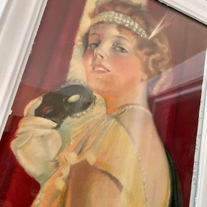 May include: A vintage framed print of a woman wearing a pearl headband and a masquerade mask. She is dressed in a yellow gown with a black shawl. The background is a red curtain.