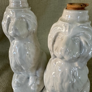 May include: Two white ceramic dog-shaped bottles with cork stoppers. The bottles have a crackled glaze and are in a sitting position.
