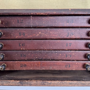 May include: A vintage wooden cabinet with five drawers, each labeled with a number. The cabinet is stained a dark reddish-brown color, with black knobs on each drawer. The numbers are painted in black. The cabinet has a rustic, antique appearance.