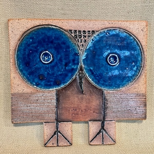 May include: A ceramic owl wall hanging with blue eyes and brown and white details. The owl has a simple, geometric design.