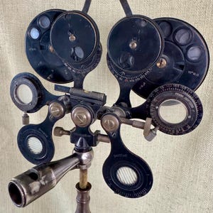 May include: An antique black ophthalmic phoropter, a device used to determine eyeglass prescriptions. It features multiple lenses, dials, and a prism, mounted on a brass and wooden stand. The device has various measurement markings.