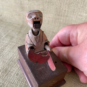 May include: A wooden toy figure of a person with a red and white painted hat, holding a red and white painted object. The figure is sitting on a wooden base.