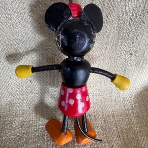 Puede incluir: Una figura de madera vintage de Mickey Mouse. La figura negra tiene manos con guantes amarillos, pies naranjas y una falda roja con lunares. Las orejas y la cabeza son negras, y el sombrero es rojo. La figura est&aacute; sobre una superficie de color neutro.