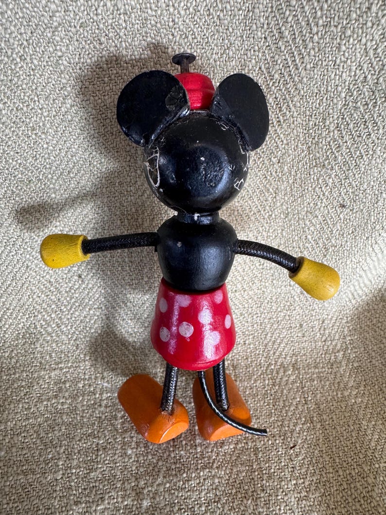 Puede incluir: Una figura de madera vintage de Mickey Mouse. La figura negra tiene manos con guantes amarillos, pies naranjas y una falda roja con lunares. Las orejas y la cabeza son negras, y el sombrero es rojo. La figura est&aacute; sobre una superficie de color neutro.