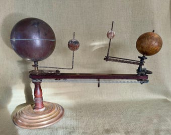 May include: A vintage wooden and metal orrery model of the solar system with three globes. The largest globe is dark brown and the other two are smaller and lighter in color. The orrery is on a wooden base with a brass ring.