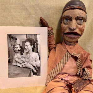 May include: A vintage wooden puppet with a painted face and a red and black striped outfit. The puppet is holding a framed black and white photograph of a woman holding a smaller puppet. The photograph is in a pink mat.