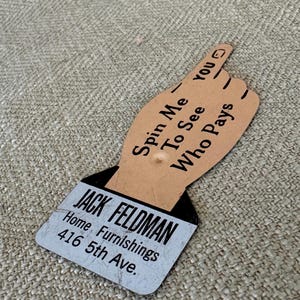 May include: A vintage, brown, cardboard hand pointing to the right with the text "Spin Me To See Who Pays" and the text "JACK FELDMAN Home Furnishings 416 5th Ave."